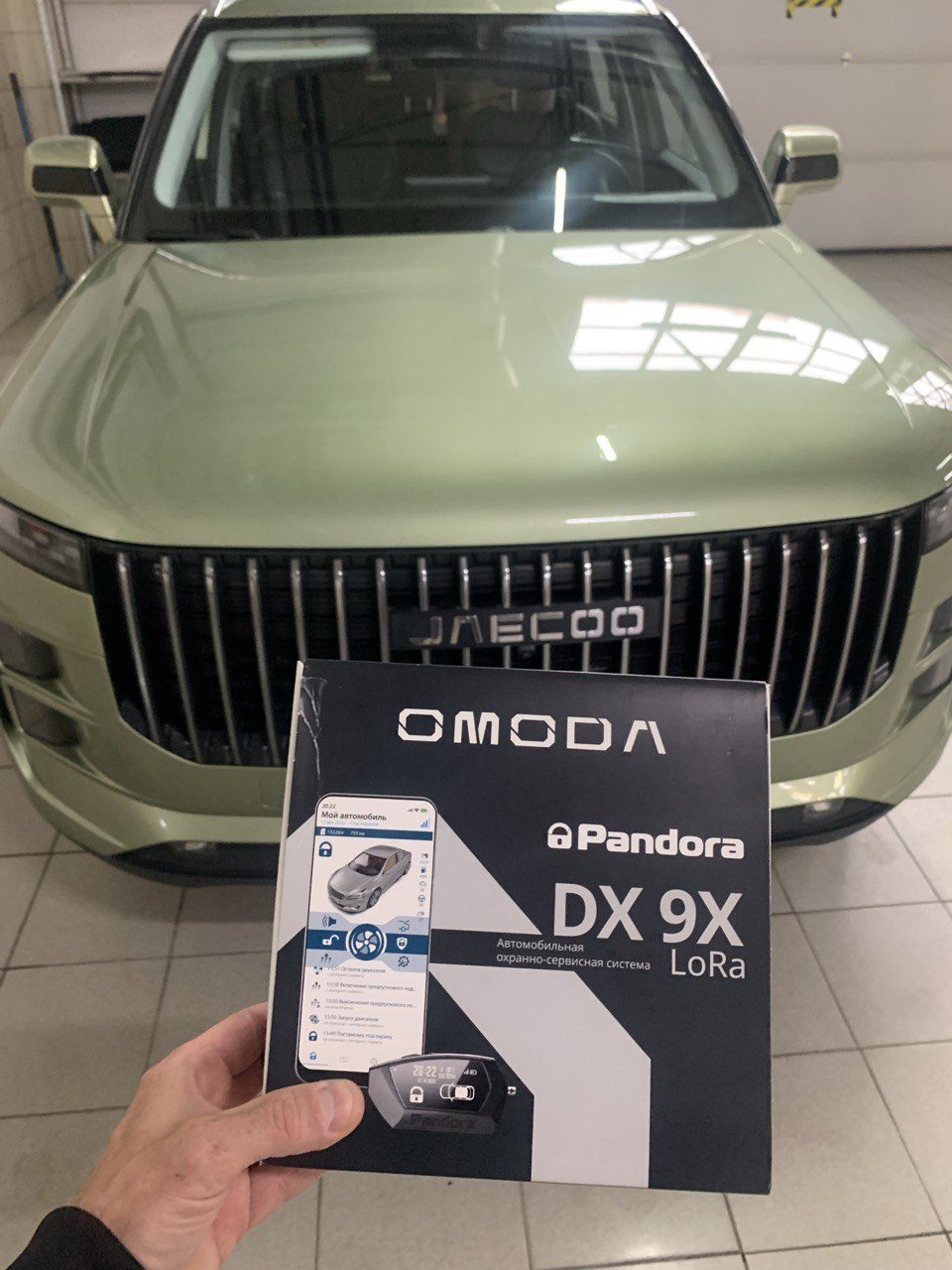 Упаковка Pandora DX 9X LoRa в руках на фоне зелёного кроссовера Jaecoo