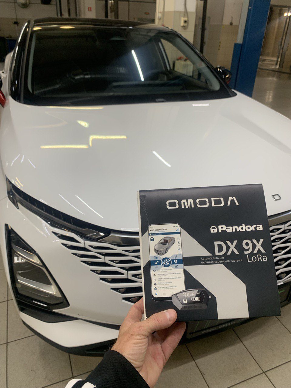 Упаковка Pandora DX 9X LoRa в руках на фоне белого кроссовера OMODA